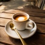 White ceramic cup of cappuccino on a saucer with a gold spoon, on a sunlit wooden outdoor table.