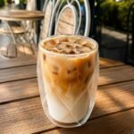 Iced latte in a double‑walled glass on a wooden patio table at an outdoor cafe, with metal chairs in the background.