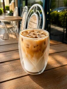 Iced latte in a double‑walled glass on a wooden patio table at an outdoor cafe, with metal chairs in the background.