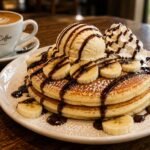 Three fluffy pancakes topped with fresh banana, chocolate sauce, scoop of ice cream and powdered sugar,