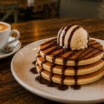 Stack of fluffy pancakes topped with vanilla ice cream, rich chocolate sauce, and whipped cream.