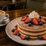 Fluffy stack of mixed berries pancakes with fresh strawberries, blueberries, powdered sugar, and maple syrup, served at Byte & Coffee.